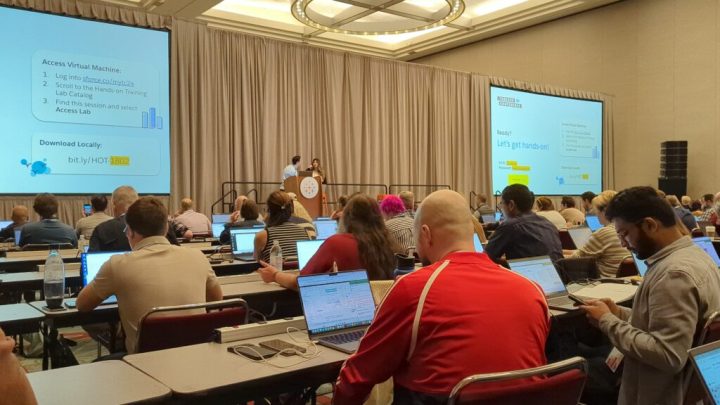 A large conference room with two screens the size of walls in a home and several rows of small tables where attendees sit and use their laptops to study the content.