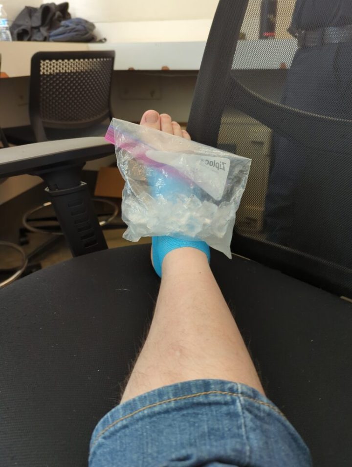 A light skin colored, somewhat hairy, right leg with jeans pulled up to just under the knee. The right foot is wrapped in light blue tape and has a Ziploc sandwich bag with ice. The foot is resting on top of an office chair