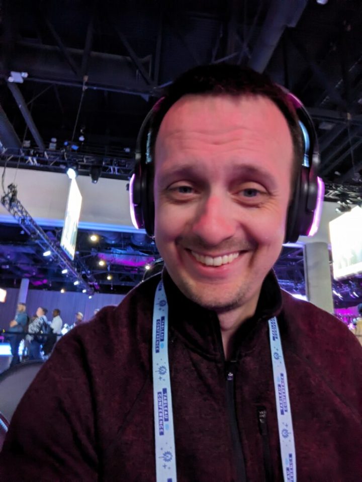 A dark-haired, blue-eyed, forty-something, white male with a thin mustache and beard facial hair combination. He is looking directly at the camera, smiling, and wearing large headphones the sides of which are colored neon pink. We only see him from the chest up. The person is wearing a half-zip, dark red sweater, and the light blue, branded Tableau Conference lanyard.