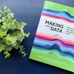 A colorful book sits on a blue table next to a small plant. The book’s cover shows a visualization made of many different colors of paper showing distribution curves. About a third of the way down the cover is the title “Making with Data Physical Design and Craft in a Data-Driven World.”