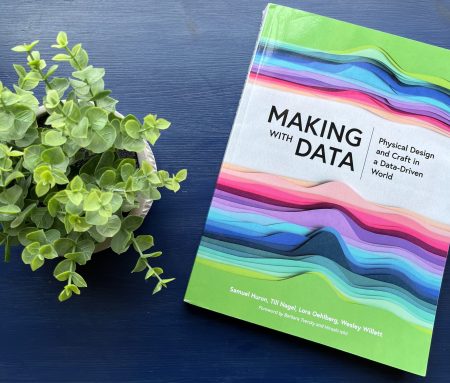 A colorful book sits on a blue table next to a small plant. The book’s cover shows a visualization made of many different colors of paper showing distribution curves. About a third of the way down the cover is the title “Making with Data Physical Design and Craft in a Data-Driven World.”