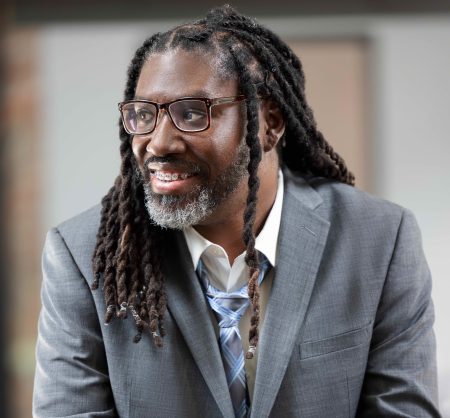 Photograph of the interview subject Larry Gray who is looking to his right side, holding his hands over a railing, and wearing glasses, a long hairstyle, and a gray suit with a loosened tie.