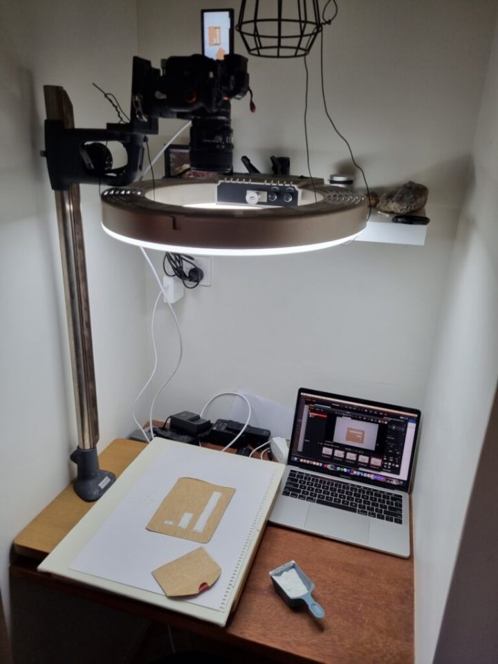 A small work space, showing an enlarger stand with a camera and light mounted on it. The camera faces down towards a piece of cardboard with mounds of washing detergent arranged into a column chart. The camera is attached to a laptop, which shows image capture software with the camera view.