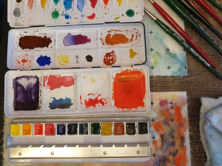 Watercolour and brushes: Photo of the messy process of using watercolours. The watercolour palette shows all the paints used in their pans and the mixes used to create the colours applied to the paper, beside the brushes used. There are also some of the many bamboo rags that contained all the paint mess.