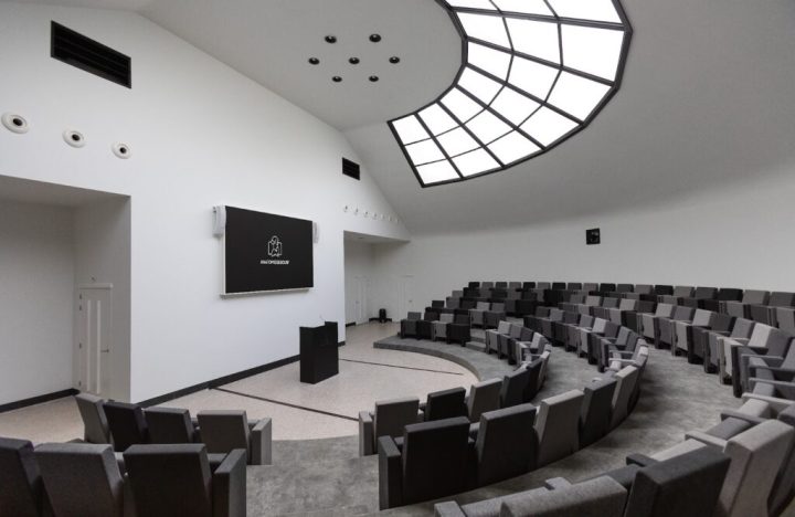 A photo of the conference room, where seats are in a semi-circle around a lectern and a screen.