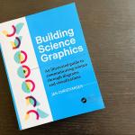 The Building Science Graphics book on a dark brown wood table. The book has a cover that is two-thirds blue and one-third white. On the white section of the cover, a series of overlapping half circles in yellow, pink, and blue are shown in varying degrees of rotation in a vertical arrangement.