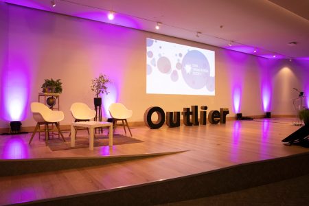 A photo of the Outlier presentation stage in Porto, with a projector screen three chairs, a podium and an physical "Outlier" letter blocks set on the floor.