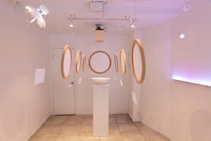 A photo of a narrow gallery space with white walls and a tiled floor. Data visuals, in circular frames, hang from the ceiling and the bowl of water on the pedestal is in the center.