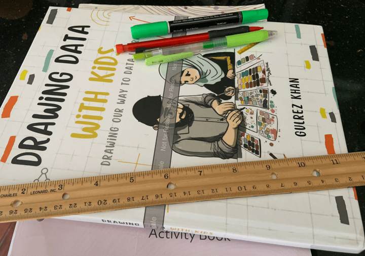Copy of the book Drawing Data With Kids covered by a ruler and several pens and pencils
