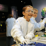 Two young boys wearing white lab coats, gloves, and safety goggles working with scientific equipment that is part of a museum exhibit