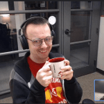 This image shows a virtual mentorship meeting between two individuals. The main focus is on a man wearing headphones and glasses, smiling warmly while holding a mug with both hands. In the bottom right corner, a smaller video frame shows a woman, his mentee, with her eyes closed and a slight smile on her face, engaged in the conversation. The atmosphere seems relaxed and friendly, suggesting a positive and comfortable mentoring relationship.