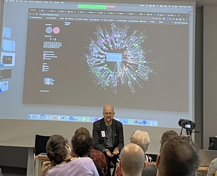 A man sits in front of a large screen providing a lecture. The screen has a projection of a network map.