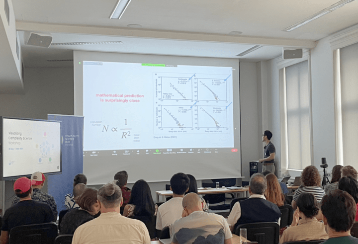 A man sits in front of a large screen providing a lecture. The screen has a projection of a scatter plots with the text "mathematical prediction is surprisingly close".